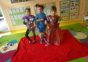 Troje dzieci w ekologicznych strojach stoi na czerwonym dywanie.